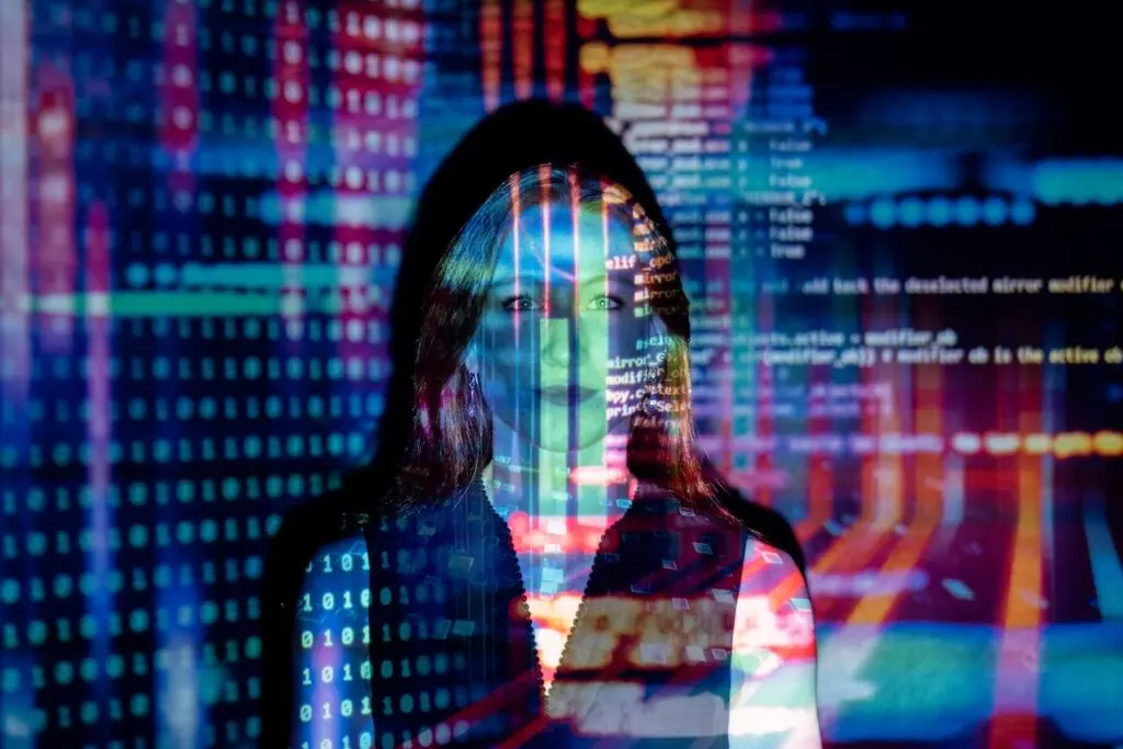A woman stands smiling in a dark room with lines of code projected over her